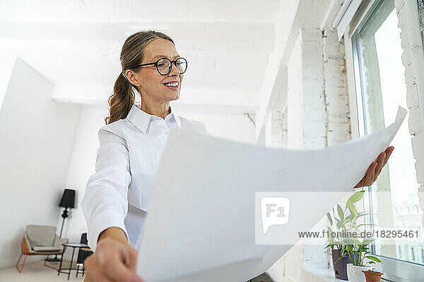 Happy businesswoman wearing eyeglasses looking at blueprint in office