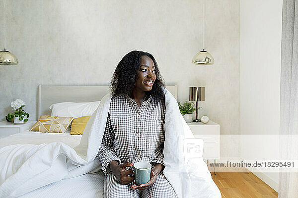 Happy young woman holding coffee cup on bed at home