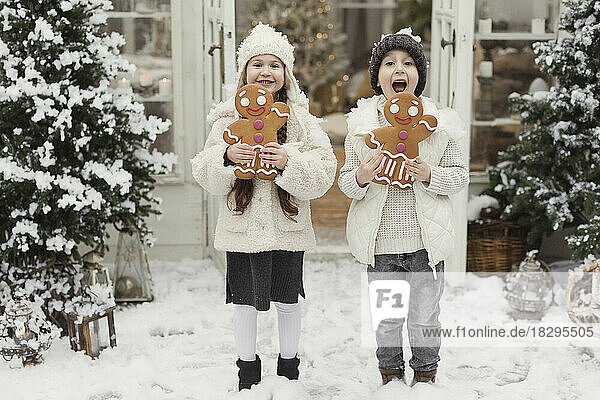 Happy siblings standing with gingerbread man cookies outside house