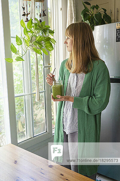 Blond woman with green smoothie looking out through window