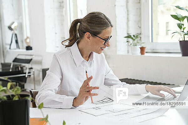 Mature businesswoman using laptop with blueprint in office