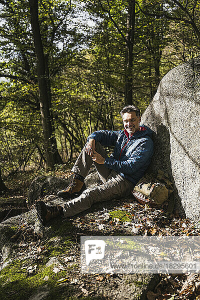 Smiling man with backpack sitting by rock in forest