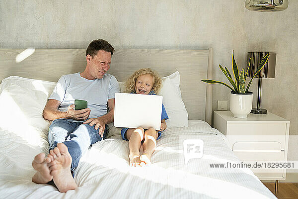 Smiling man with mobile phone looking at daughter using laptop on bed