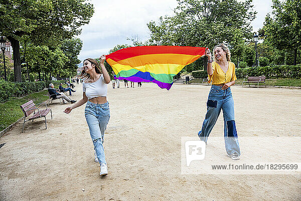 Smiling lesbian young couple holding rainbow flag walking in park