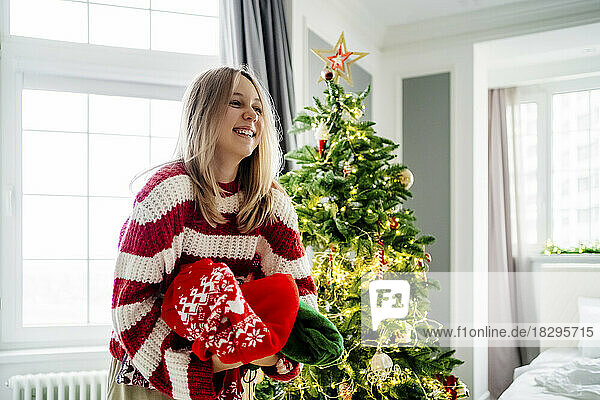Cheerful girl holding sweaters by Christmas tree at home