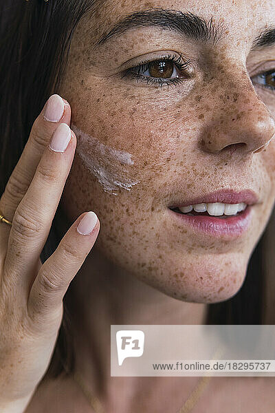 Young woman with freckle face applying moisturizer on face