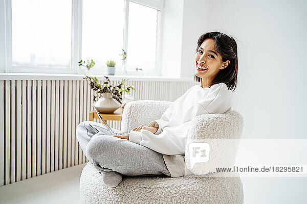 Happy young woman with laptop sitting on chair at home