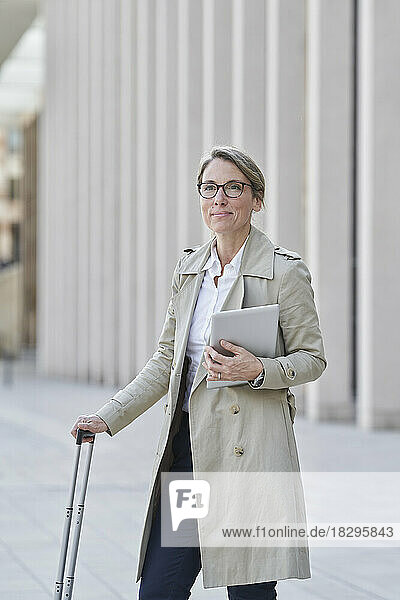 Reife Geschäftsfrau mit Rollgepäck steht in der Stadt und hält einen Tablet-PC in der Hand