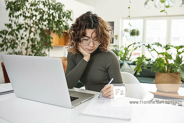 Freelancer writing notes sitting in front of laptop at home