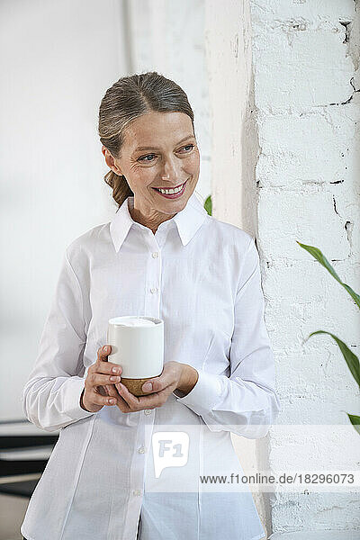 Happy mature businesswoman with coffee cup standing by wall