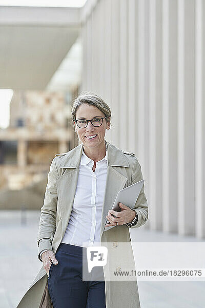 Confident businesswoman holding tablet PC standing with hand in pockets