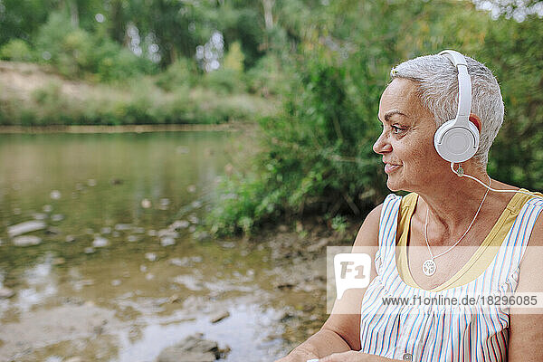 Senior woman wearing headphones listening to music by lake in park