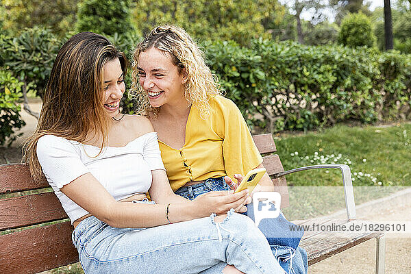 Happy woman sharing mobile phone with friend on bench in park