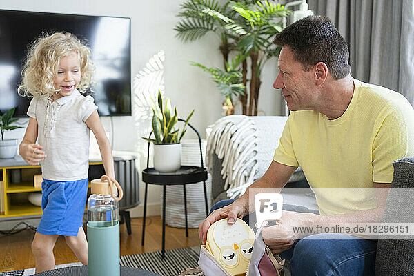 Father putting lunch box in bag and looking at daughter in living room