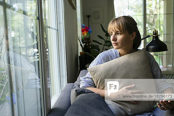 Nachdenkliche Frau mit Kissen sitzt zu Hause auf dem Sofa