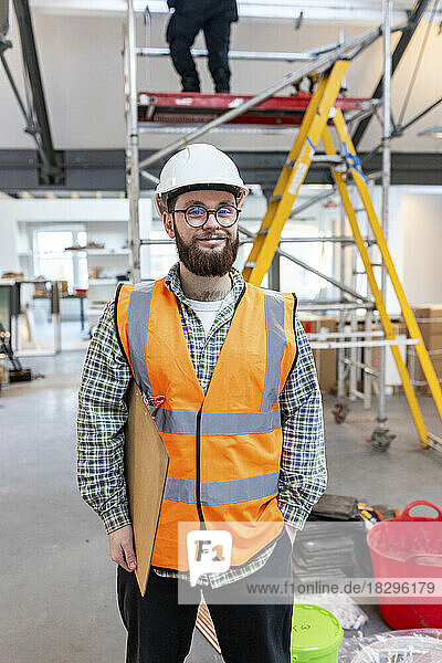 Smiling young engineer holding clipboard standing at construction site
