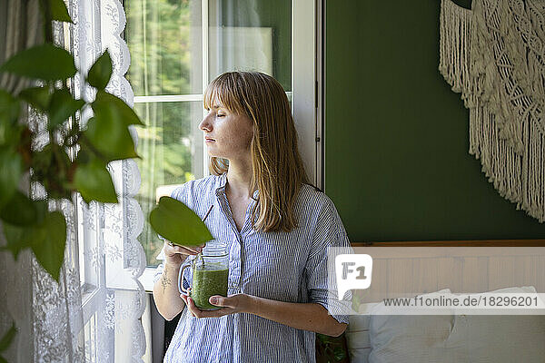Woman with eyes closed holding smoothie standing by window in bedroom