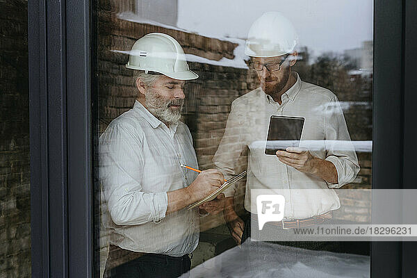 Architects wearing hardhat discussing using tablet PC seen through glass
