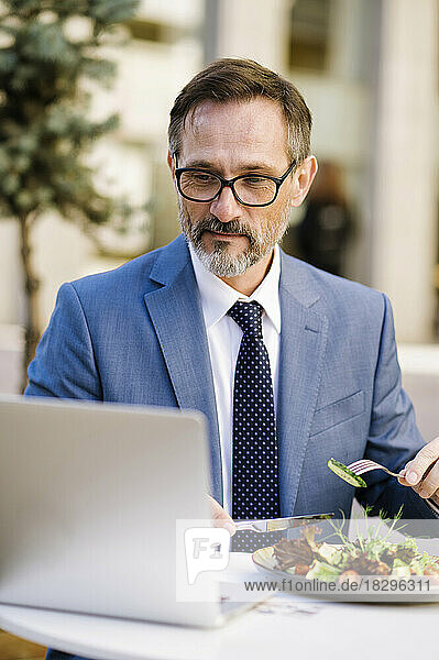 Mature businessman working and eating lunch in front of laptop at cafe