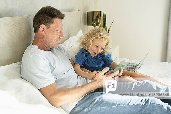 Father sharing mobile phone with daughter on bed in bedroom