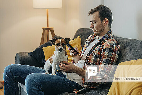 Mann benutzt Smartphone und sitzt mit Hund zu Hause auf dem Sofa