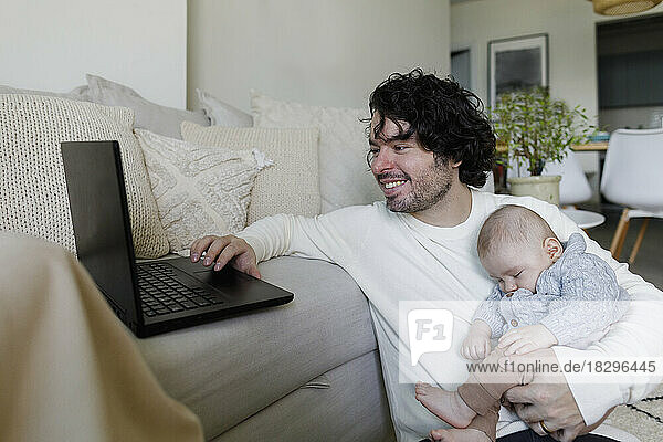 Happy freelancer working on laptop with baby boy sleeping at home