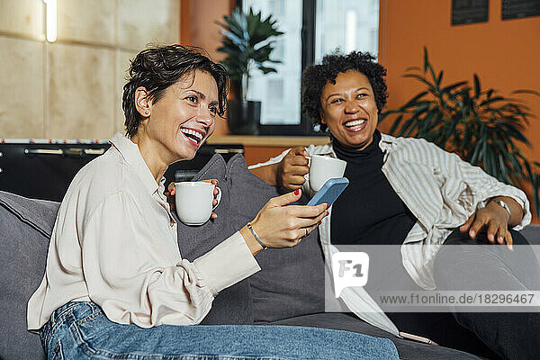 Happy businesswomen with coffee cups sitting on sofa in office