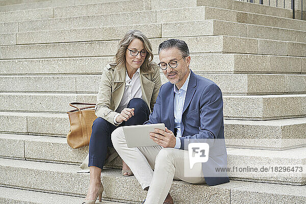 Mature businessman using tablet PC sitting by colleague on steps
