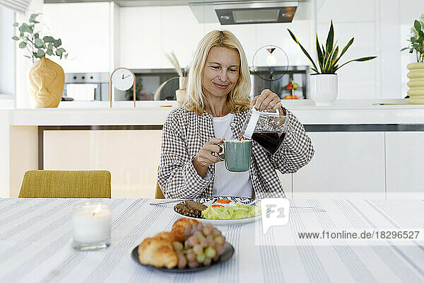 Lächelnde Frau  die am Esstisch sitzt und Kaffee in eine Tasse gießt