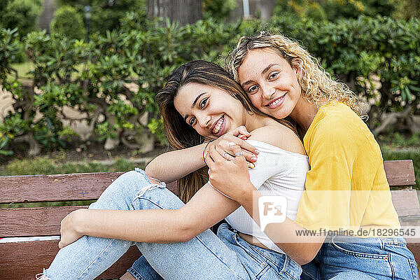 Smiling woman with blond hair embracing girlfriend from behind on bench in park