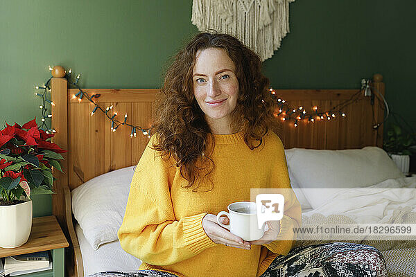 Smiling woman holding cup of tea on bed at home