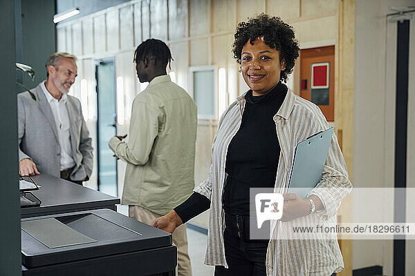 Smiling businesswoman standing by photocopier machine with colleagues standing in background