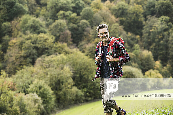 Lächelnder Mann mit Rucksack wandert auf Gras vor Bäumen