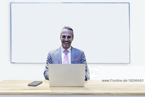 Smiling businessman working on laptop sitting in front of whiteboard at office