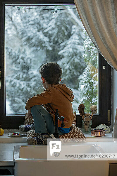 Boy sitting on window sill looking out in kitchen at home