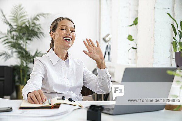Glückliche reife Geschäftsfrau winkt mit Laptop auf dem Schreibtisch im Büro
