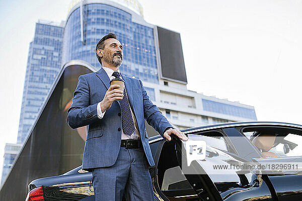 Contemplative businessman with disposable coffee cup standing by car outside building