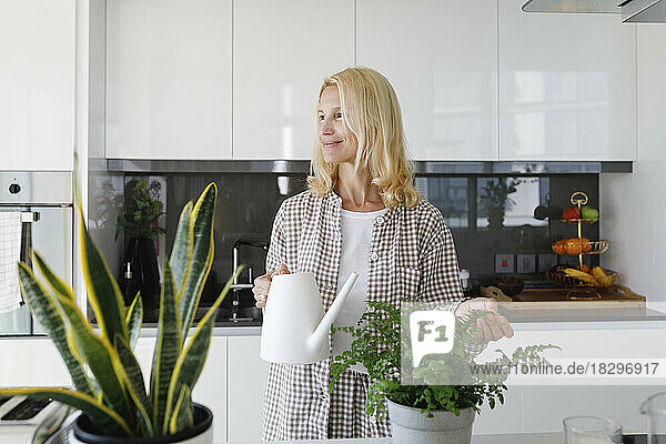 Lächelnde Frau mit Gießkanne steht zu Hause in der Küche neben einer Topfpflanze