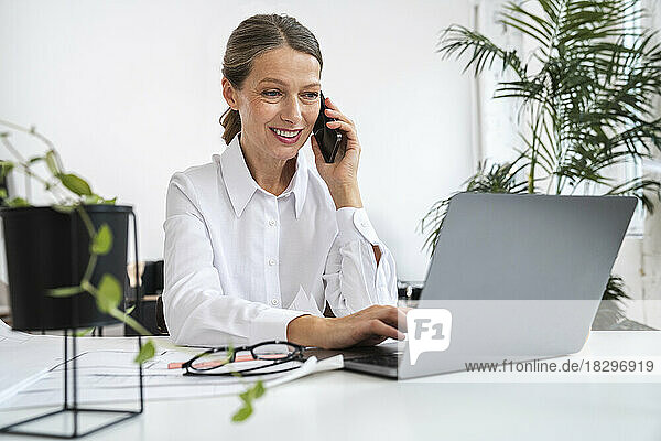 Lächelnde Geschäftsfrau  die im Büro mit dem Laptop telefoniert
