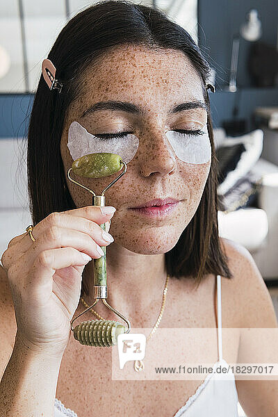 Woman with under eye patches massaging using jade stone roller on face at home