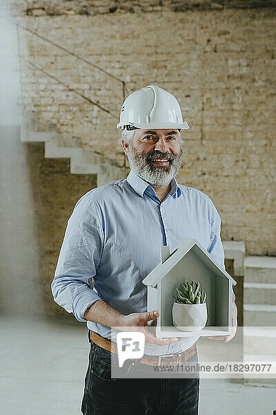 Happy businessman holding house model with potted plant at construction site
