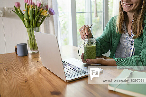 Happy freelancer with smoothie sitting at table in kitchen at home