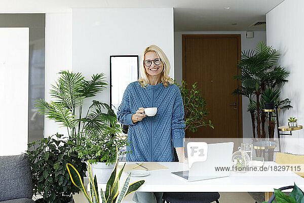 Happy freelancer standing with tea cup by desk at home