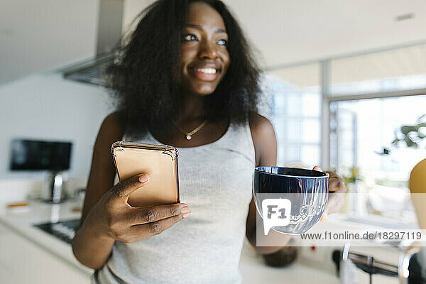 Smiling woman holding smart phone and tea cup at home