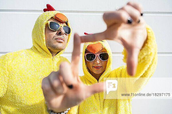 Friends wearing yellow chicken costumes looking through finger frame in front of wall