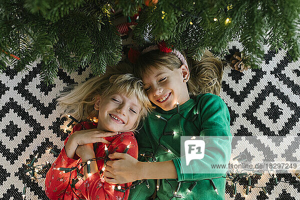 Happy sisters with illuminated string lights lying down near Christmas tree