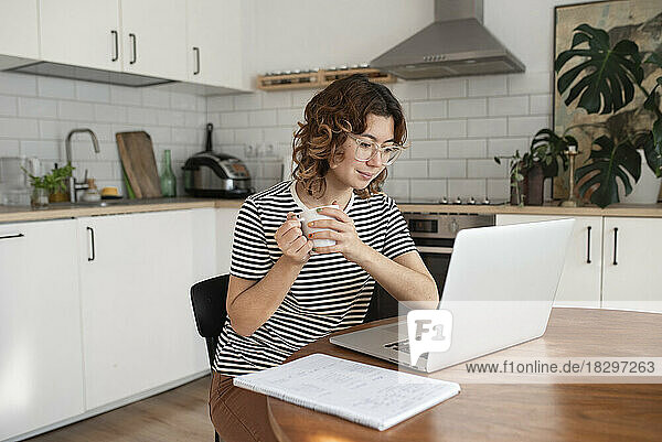 Freelancer holding coffee cup using laptop on table at home