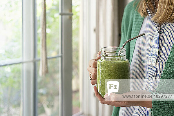 Hands of woman with green smoothie by window