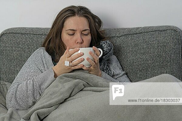 Mit einer Decke zugedeckte kranke Frau  die auf der Couch in ihrer Wohnung eine Infusion trinkt