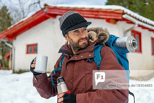 Mann trinkt im Winter im Schnee neben einer Hütte Kaffee aus einer heißen Thermoskanne  nachdem er darin geschlafen hat